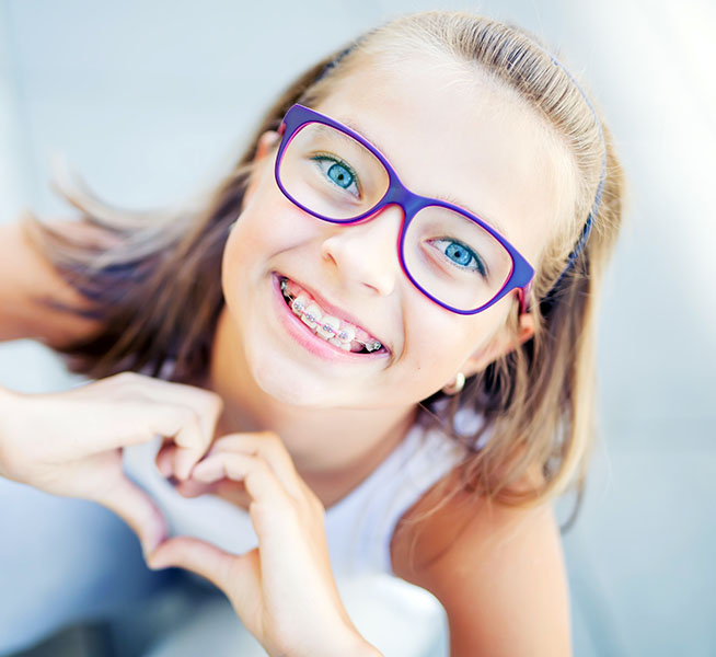 Smiling little girl in with braces and glasses showing heart with hands.